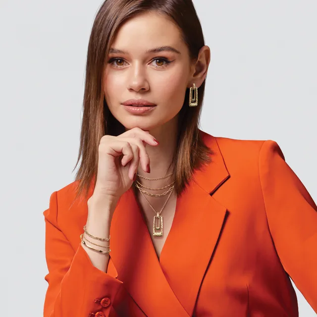 Woman wearing a bright orange blazer with gold chain necklaces and rectangular drop earrings, posed against a light gray background with her hand near her chin.