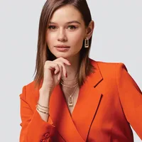 Woman wearing a bright orange blazer with gold chain necklaces and rectangular drop earrings, posed against a light gray background with her hand near her chin.