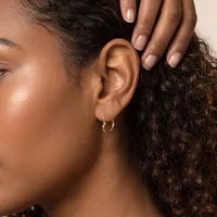 A woman with brown skin and curly dark hair wears small gold hoop earrings and displays her manicured nails while touching her ear.