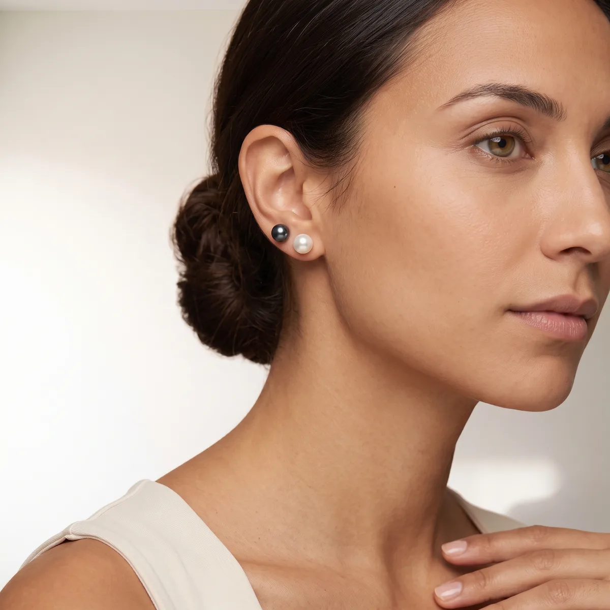 A woman in profile wearing small round pearl stud earrings and a cream-colored sleeveless top, with her dark hair styled in a low bun.