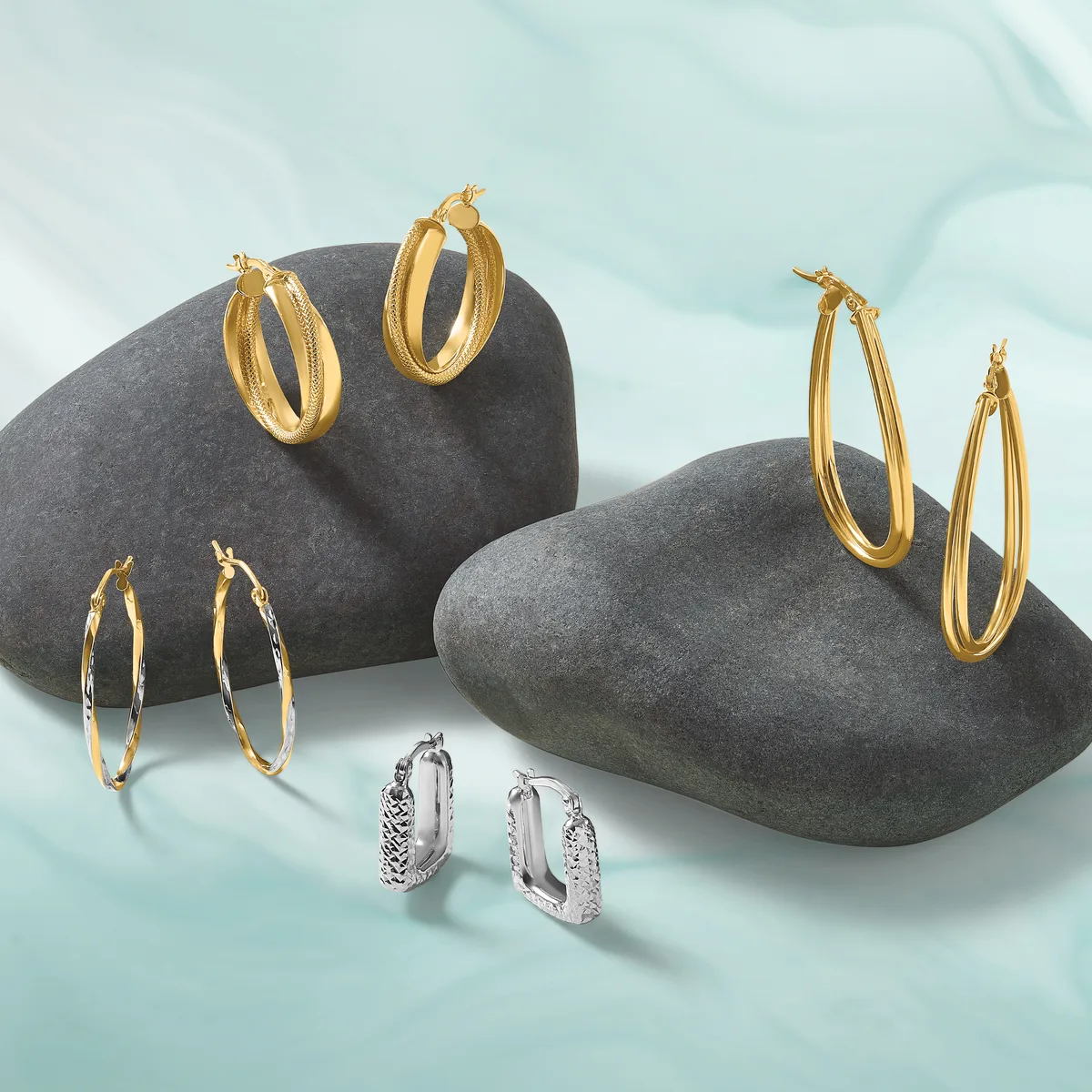 Five pairs of hoop earrings displayed on dark gray stones against a light blue background: three gold-toned pairs in various sizes and textures, and two silver-toned pairs with textured surfaces.