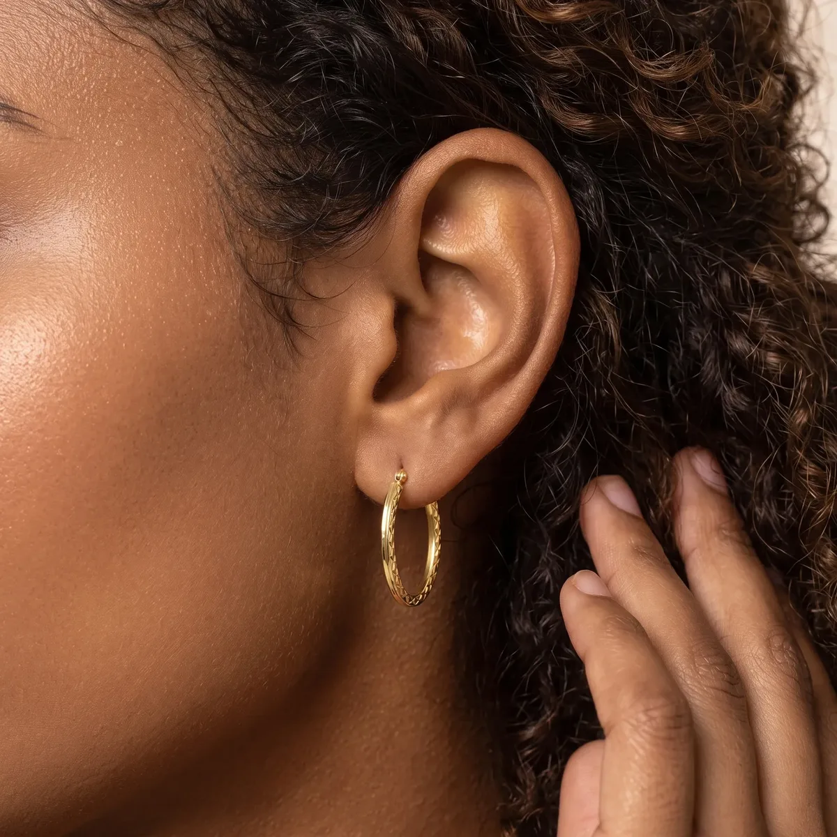 A person wearing a gold textured hoop earring is shown in profile, with curly dark hair and their hand near their face.
