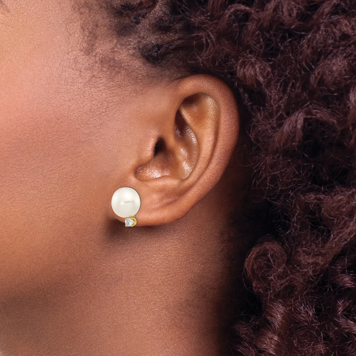 A person wearing a white pearl stud earring with a small diamond accent, shown from the side profile against their ear and dark curly hair.