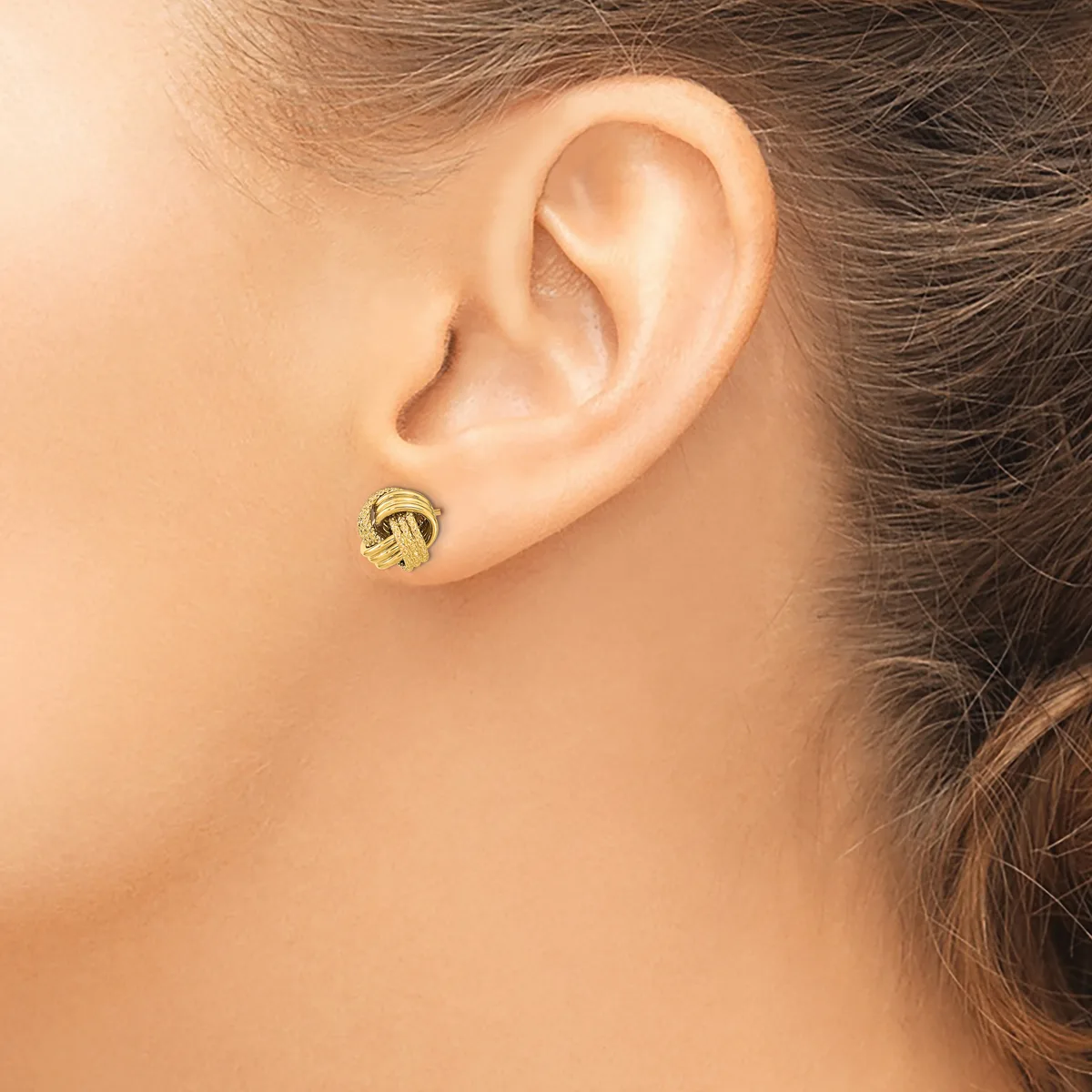 A woman wearing a small gold knotted stud earring in her earlobe, shown from the side view of her ear and neck.