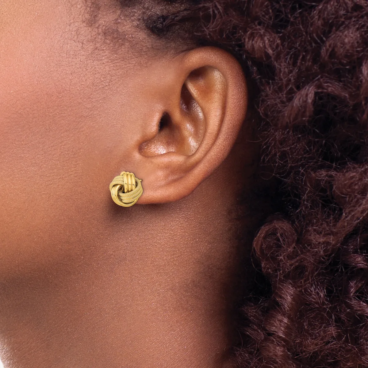 A person wearing a gold-toned twisted knot stud earring in their earlobe, shown in profile against their neck and shoulder.