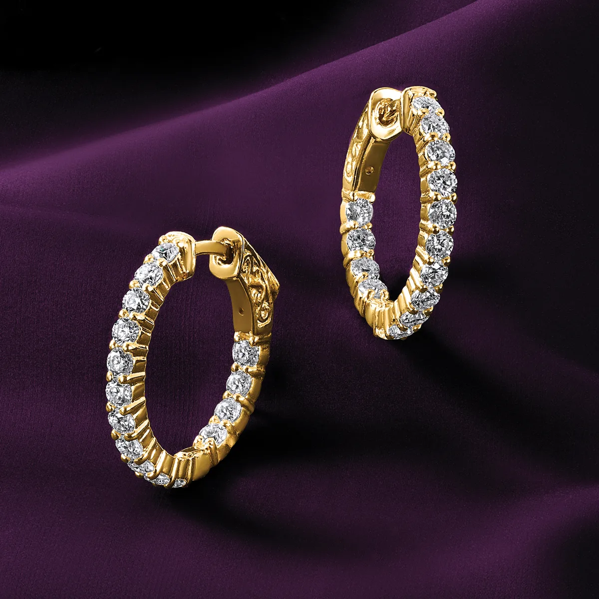Two gold hoop earrings set with round diamonds arranged in a line, displayed against a purple fabric background.