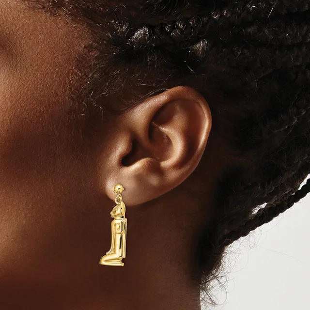 A gold-toned candle-shaped dangle earring hangs from a pierced earlobe against a person's brown skin and dark curled hair.
