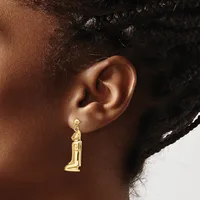 A gold-toned candle-shaped dangle earring hangs from a pierced earlobe against a person's brown skin and dark curled hair.
