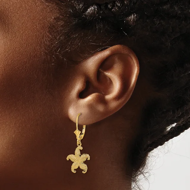 A person wearing a gold starfish-shaped pendant earring with a lever back closure, shown hanging from their earlobe against their skin.