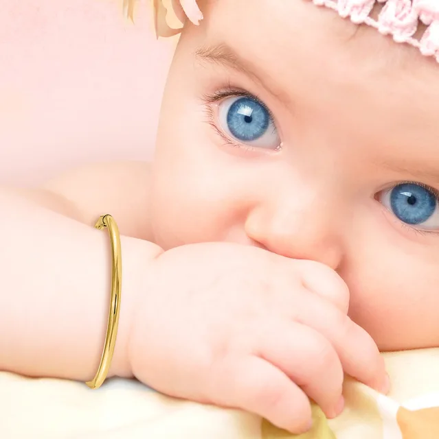A baby with bright blue eyes and blonde hair wearing a gold-toned bangle bracelet, with their hand near their mouth against a soft pink background.