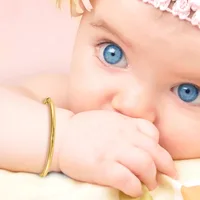A baby with bright blue eyes and blonde hair wearing a gold-toned bangle bracelet, with their hand near their mouth against a soft pink background.