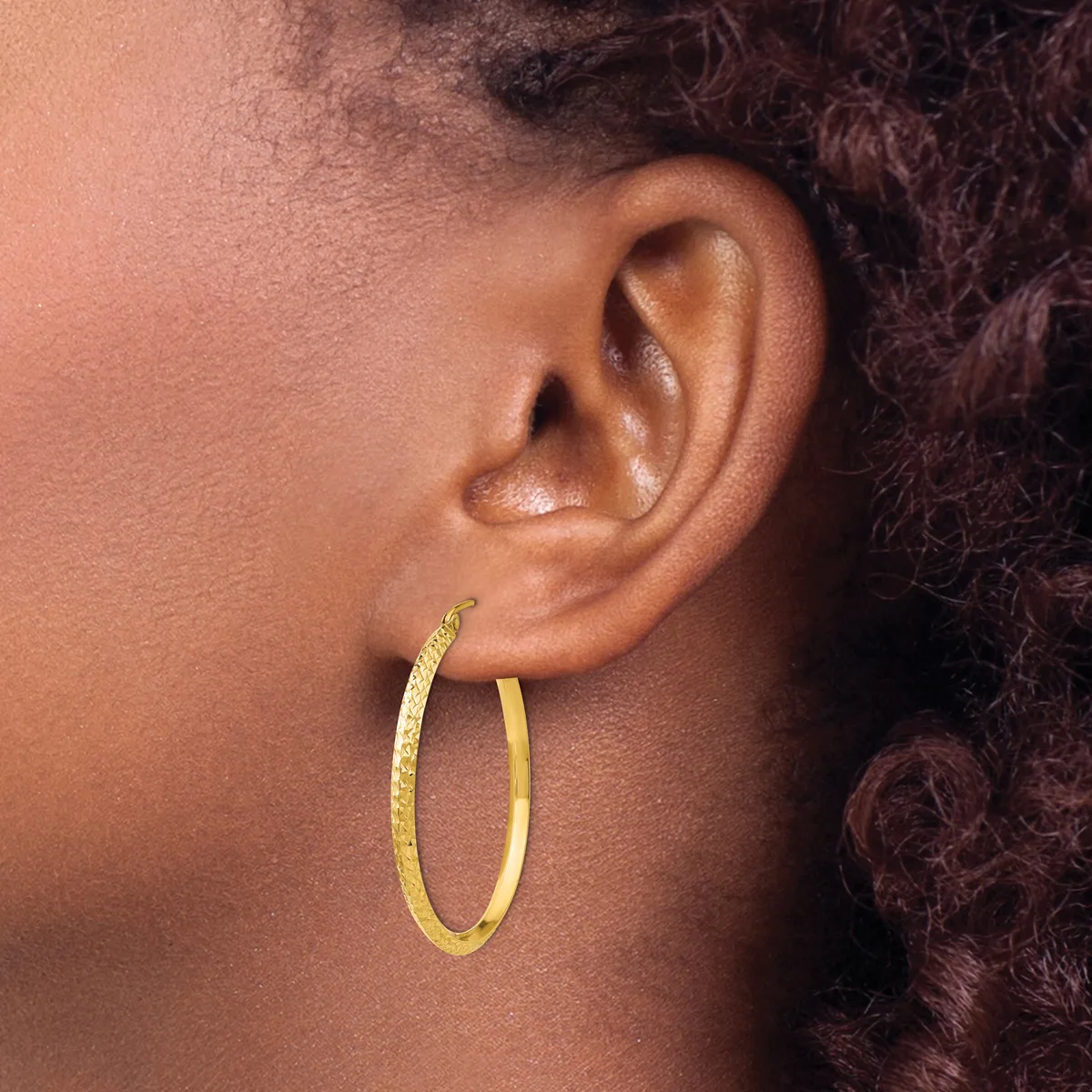 A person wearing a pair of textured gold hoop earrings, shown from the side profile with dark curly hair visible.