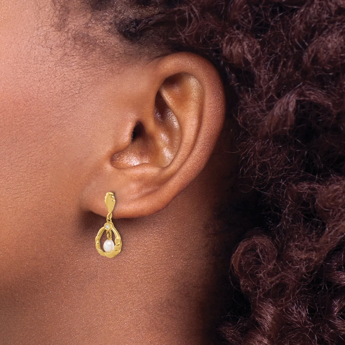 A person wearing a gold teardrop-shaped earring with a white stone or pearl center, shown from the side with dark curly hair visible.