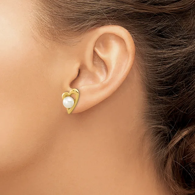 A person wears a gold-toned heart-shaped earring with a white pearl center in their earlobe.