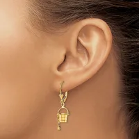 A person wearing a gold bucket-shaped charm earring with a handle, suspended from a gold lever-back hook, shown from the side view of their ear.