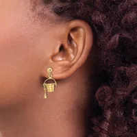 A person wearing gold-toned earrings featuring a small ball stud and a dangling bucket-shaped charm with a handle and small teardrop detail.