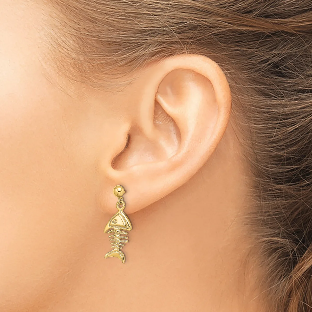 A gold fish skeleton-shaped dangle earring hanging from a small round post on a pierced earlobe.