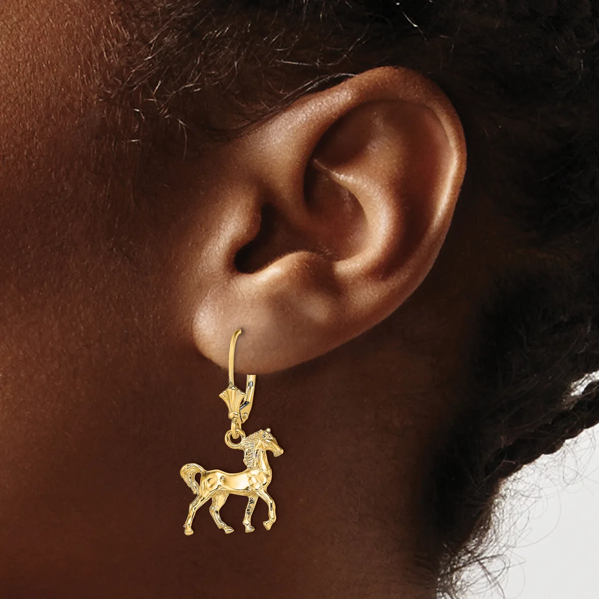 A gold horse charm earring hangs from a gold hook through a pierced ear against a dark background.