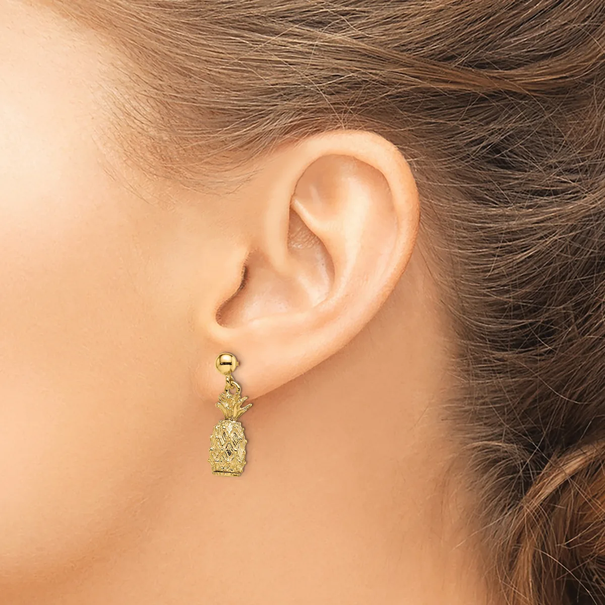 A gold pineapple-shaped drop earring hanging from a post on a woman's earlobe.