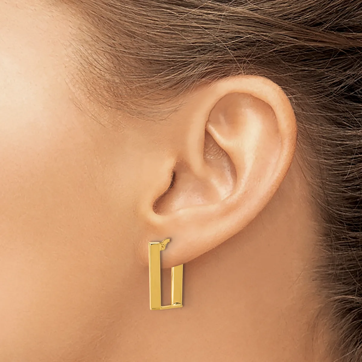A person wearing a pair of gold rectangular hoop earrings with a polished finish, shown from the side view of their ear.