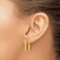 A person wearing a pair of gold rectangular hoop earrings with a polished finish, shown from the side view of their ear.