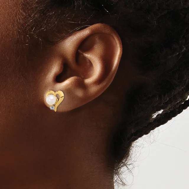 A gold heart-shaped earring with a white pearl accent on the earlobe of a person with brown skin and dark hair.