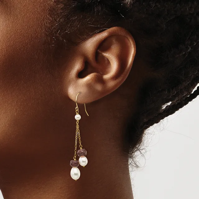 A person wearing gold chain drop earrings with white pearl and rose gold coin charms hanging from hook ear wires.