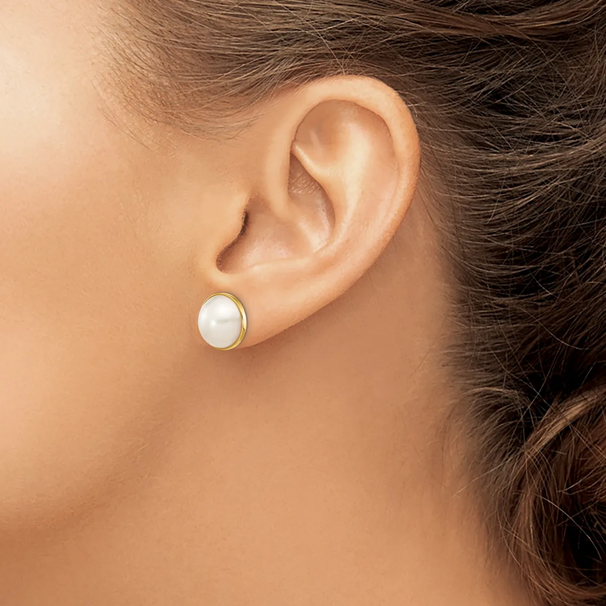A woman wearing a pearl stud earring with a gold bezel setting, shown from the side profile against her ear.