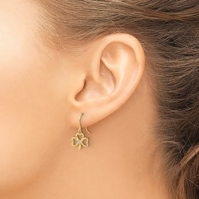 A person wears a gold four-leaf clover charm earring on a hook wire, shown from the side view of their ear and face.
