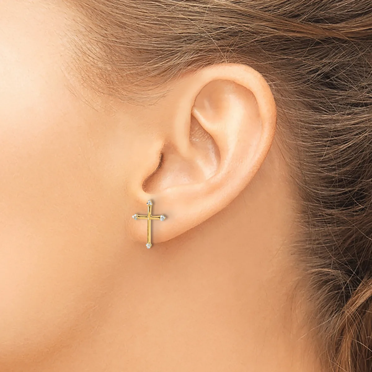 A person wearing a gold cross earring with diamond accents in their pierced earlobe, shown from the side profile.