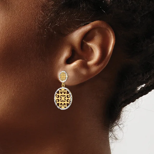 A person wearing a two-tier drop earring with a small round stud top and a larger oval pendant bottom, both featuring gold and white enamel openwork designs, shown from the side view of the ear.