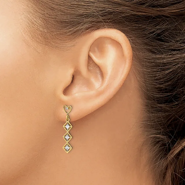 A woman wearing a long drop earring with a heart-shaped stud and three diamond-shaped gemstone links in gold-toned metal.