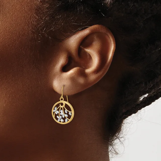 A gold-toned circular drop earring with an openwork leaf or vine pattern hangs from a wire hook on a person's ear.