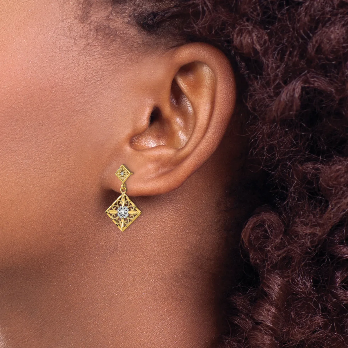 A person wearing a gold diamond-shaped drop earring with a small square stud, shown from the side profile against their neck and shoulder.