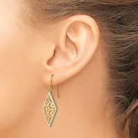 A woman wears a diamond-shaped dangle earring with filigree detailing in two-tone metal on a wire hook through her earlobe.