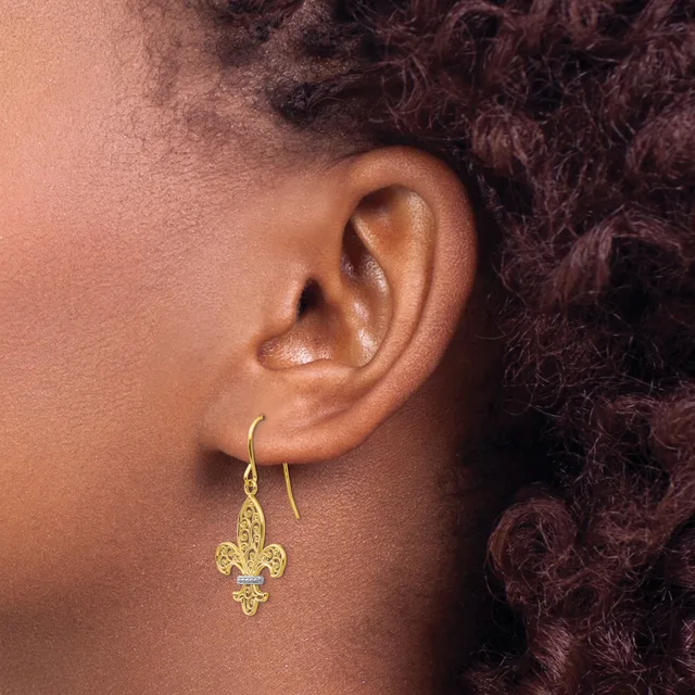 A close-up view of a person's ear wearing a gold fleur-de-lis drop earring with a small blue stone accent, shown in profile against dark curled hair.