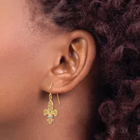 A close-up view of a person's ear wearing a gold fleur-de-lis drop earring with a small blue stone accent, shown in profile against dark curled hair.