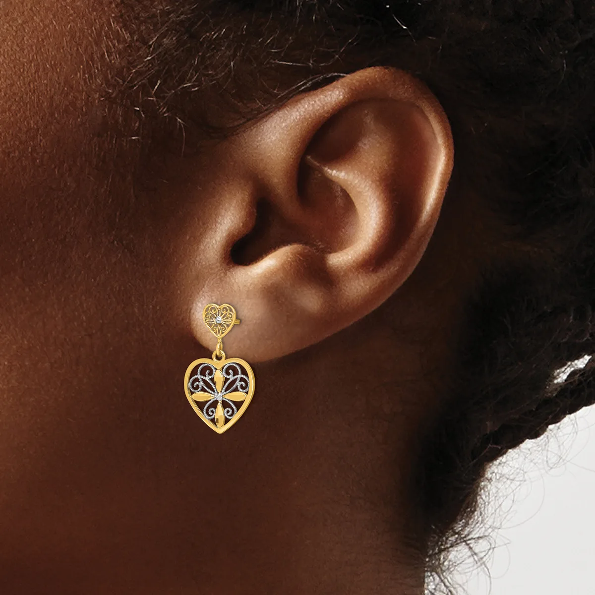 Close-up of a person's ear wearing a gold heart-shaped pendant earring with filigree detailing and a small circular stud above it.