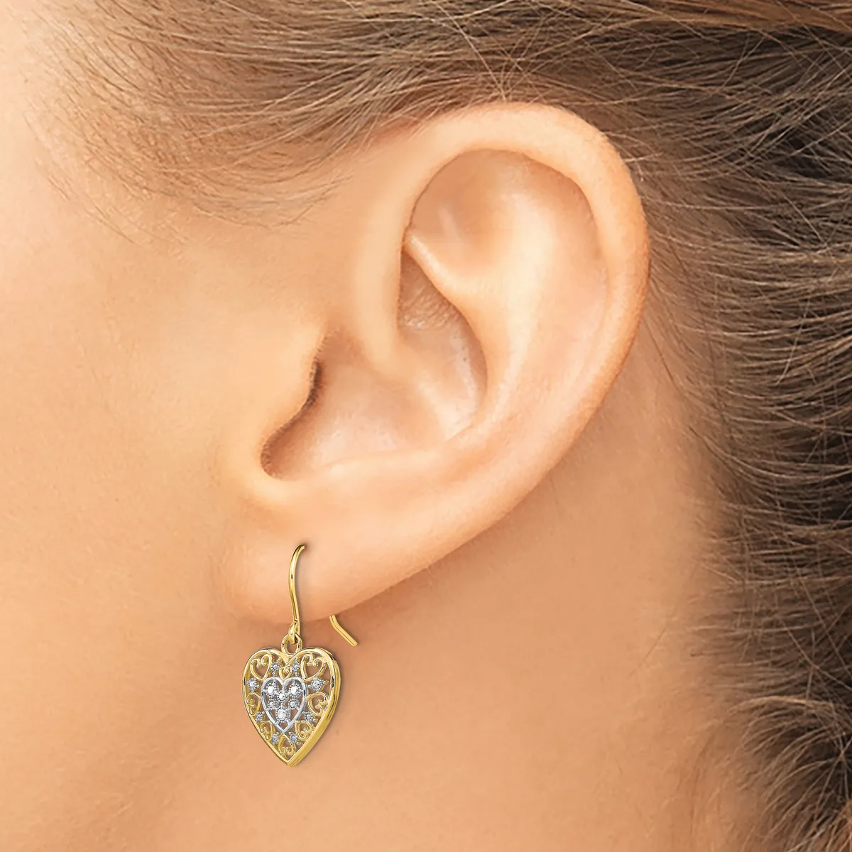 A person wearing a gold heart-shaped dangle earring with a filigree pattern and small gemstones, shown from the side view of their ear.