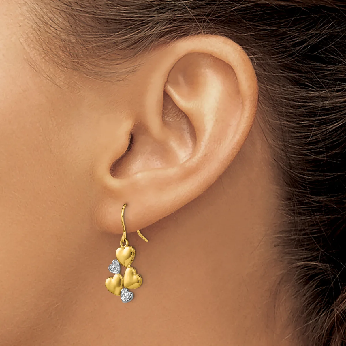 A person wearing gold drop earrings with heart-shaped charms and diamond accents, shown from the side view of their ear and neck.