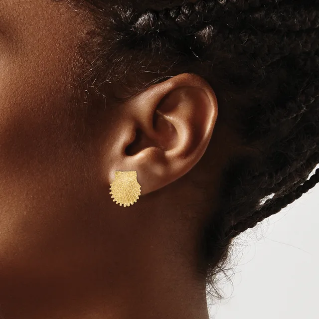 A close-up view of a person's ear wearing a small gold scallop shell-shaped stud earring with a textured finish.