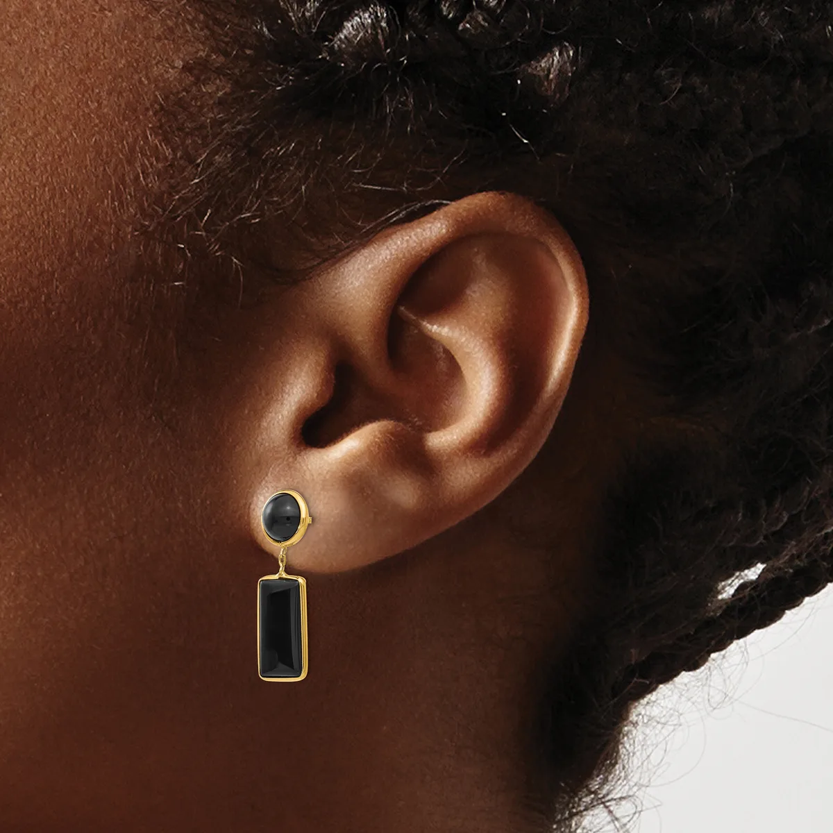 A person wearing a two-part dangling earring with a small round black stone stud on top and a rectangular black stone pendant below, both set in gold-toned metal.