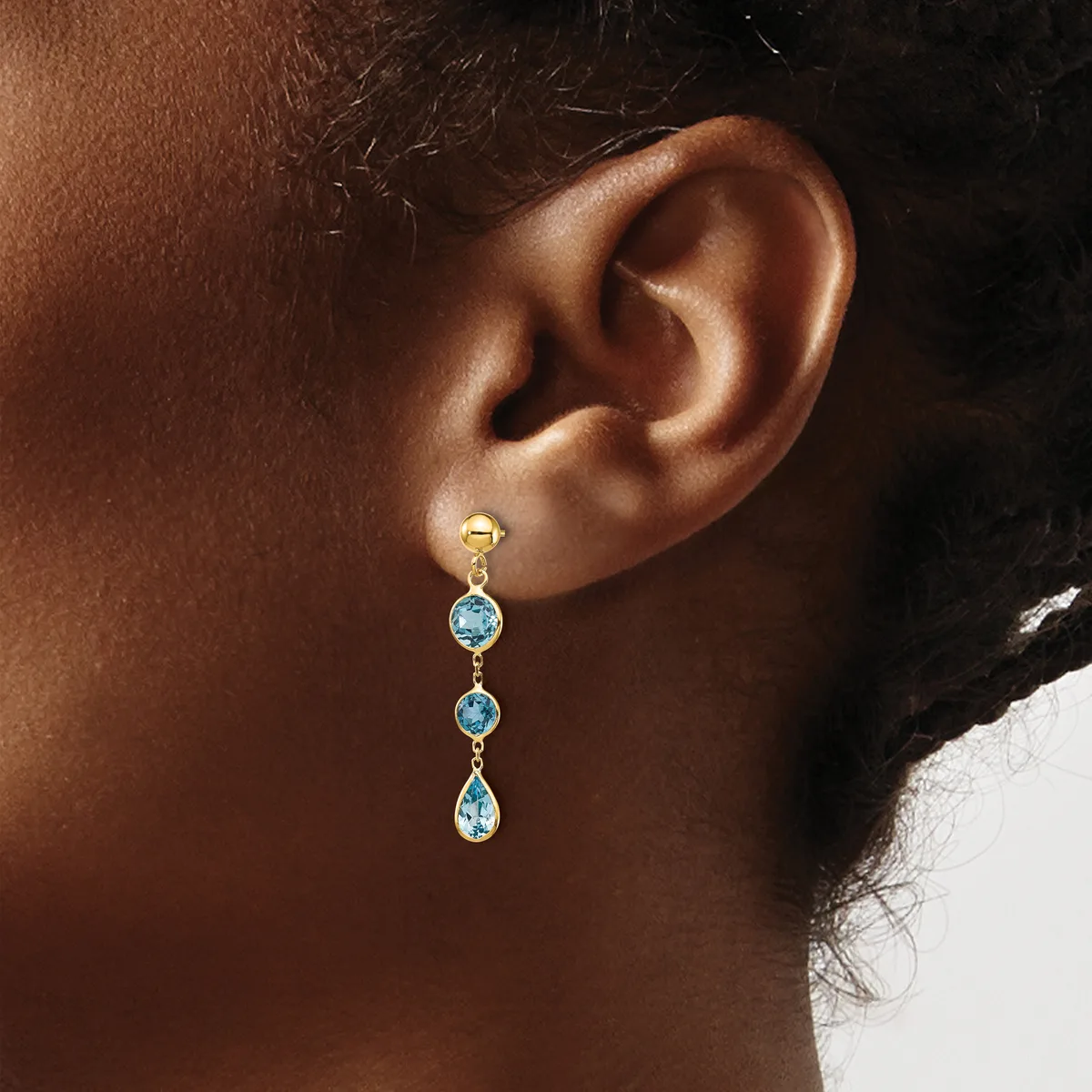 A person wearing a gold drop earring with three round blue topaz stones connected by gold links, shown from the side profile.