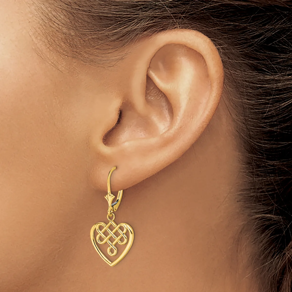 A woman wearing a gold-toned heart-shaped pendant earring with an interlocking Celtic knot design, suspended from a matching lever-back hook.
