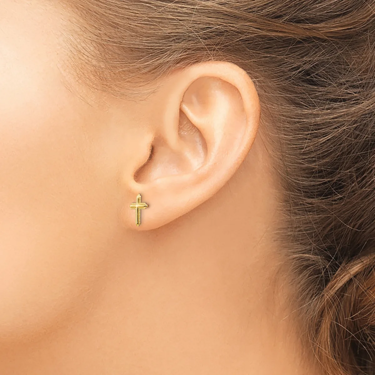 A small gold cross stud earring is worn in the earlobe of a person with brown hair.