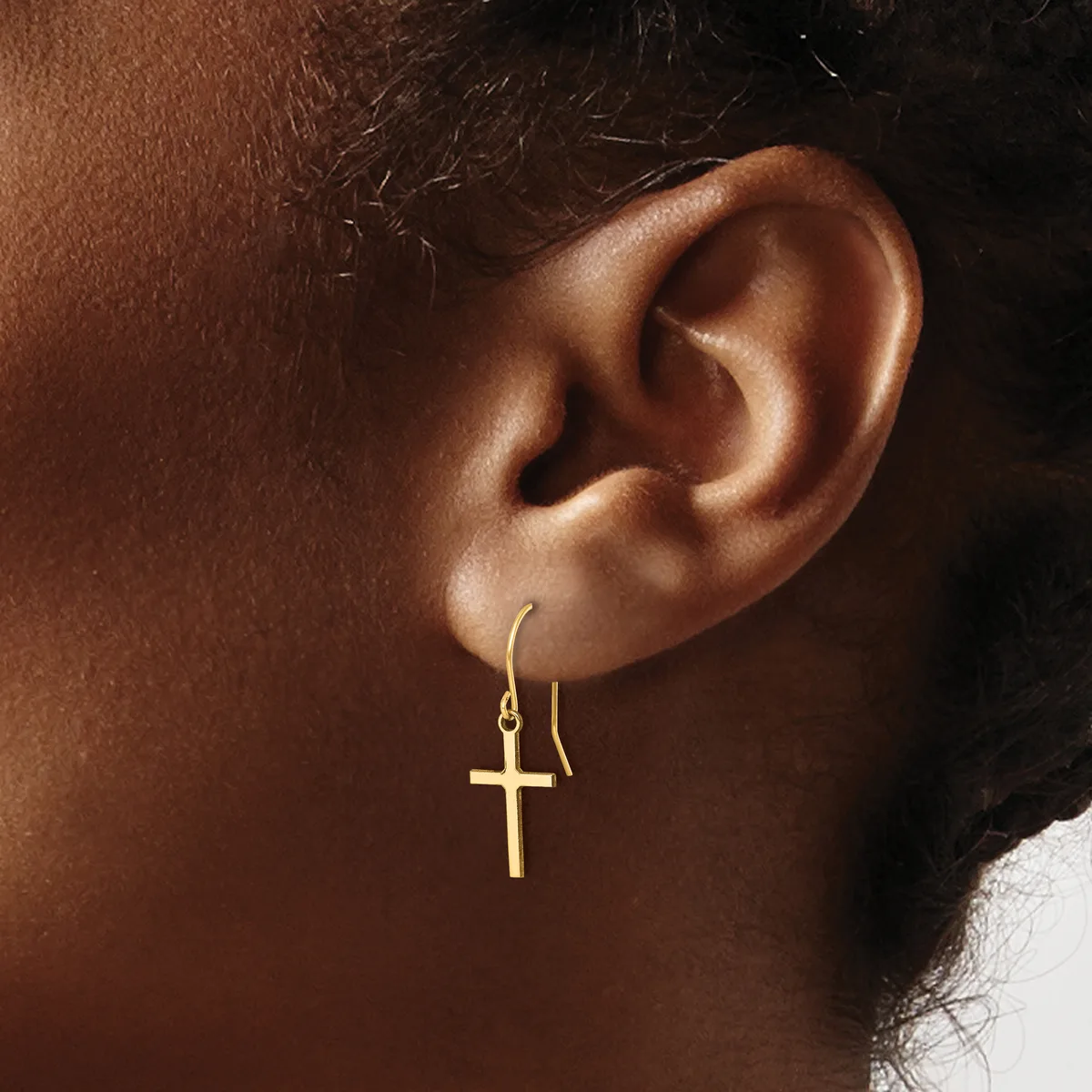 Close-up of a person's ear wearing a gold cross pendant hanging from a thin curved hook earring.