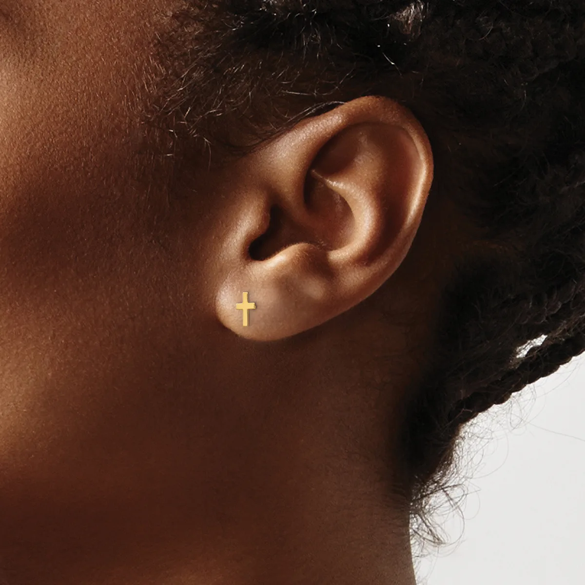 A small gold cross earring is worn on the earlobe of a person shown in profile against a white and dark background.