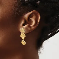 A person wearing a long gold drop earring with a textured circular disc and shell-shaped pendant, shown from the side against their ear.