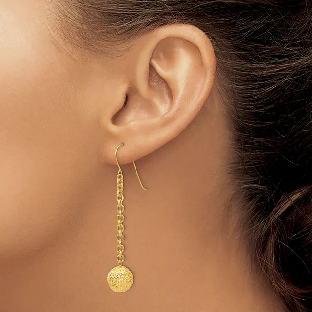 A person wearing gold drop earrings with textured spherical pendants hanging from curved wire hooks, shown from the side profile near the ear.