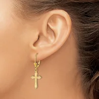 A person wearing a gold-toned cross pendant earring with textured surface detail, shown hanging from a lever-back ear wire.